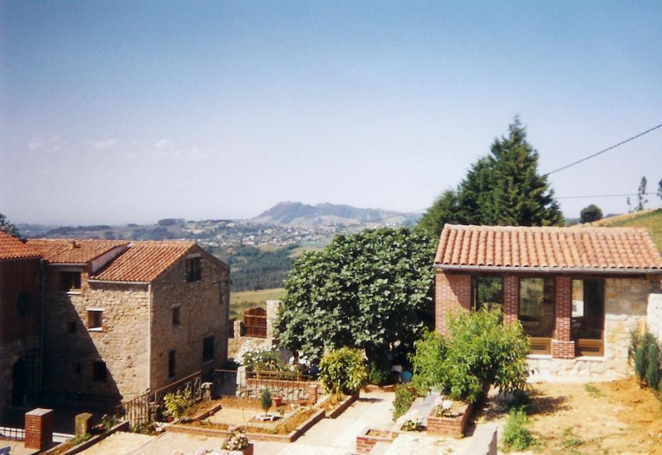 Elevated view of stone and brick buildings with terracotta roofs, garden terraces, trees, and distant hills under a clear sky, quiet and reflective mood